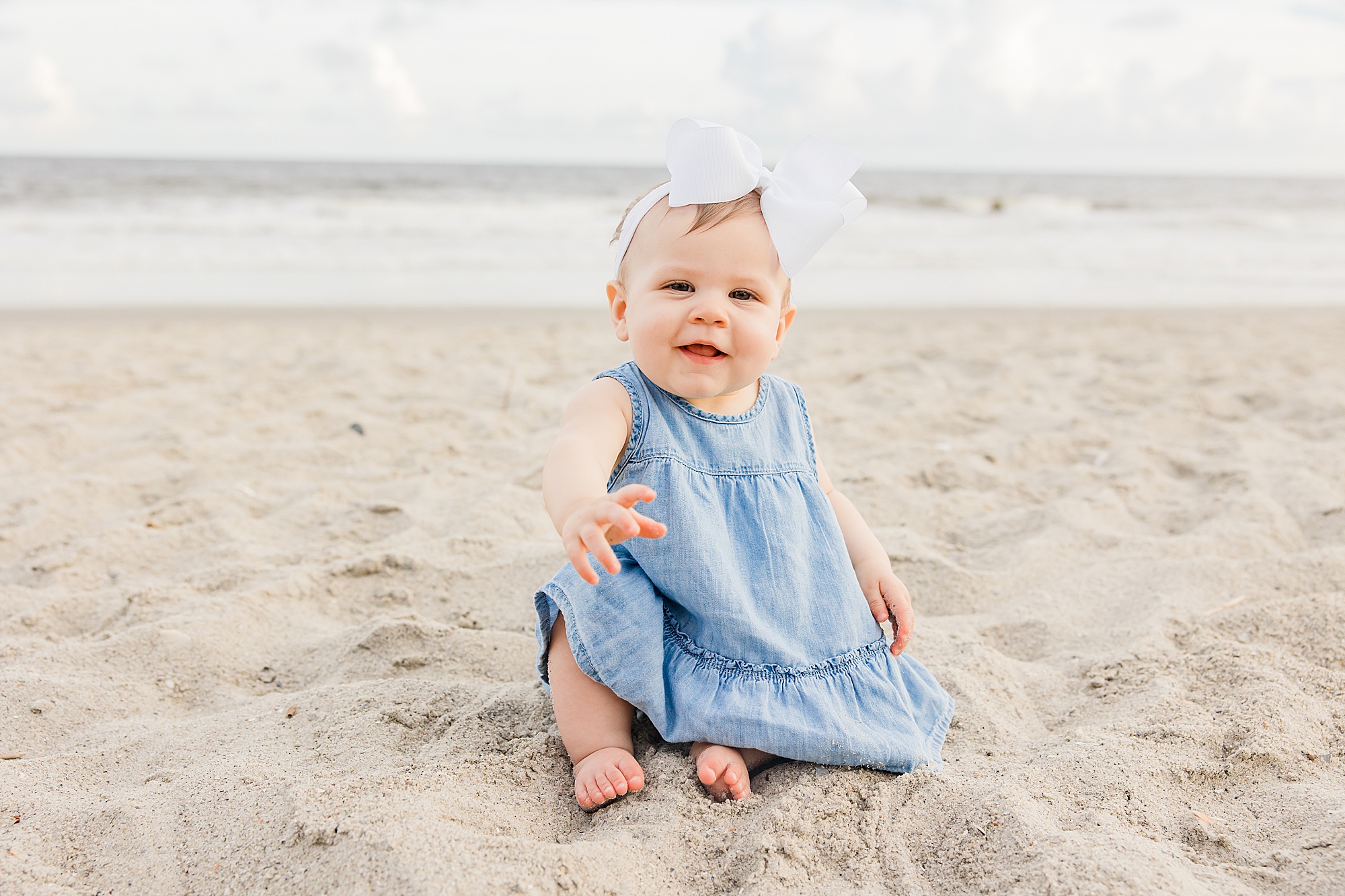 toddler in blue dress with white bow sits in sand during family photos