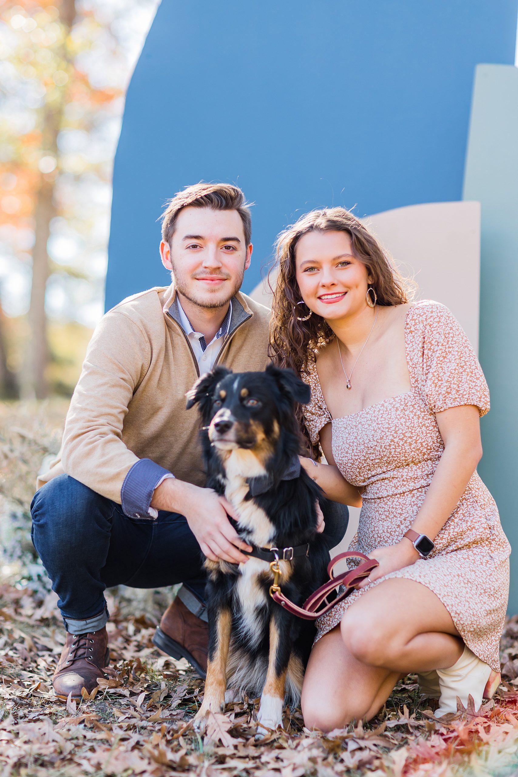 couple poses with dog by blue backdrop during mini session