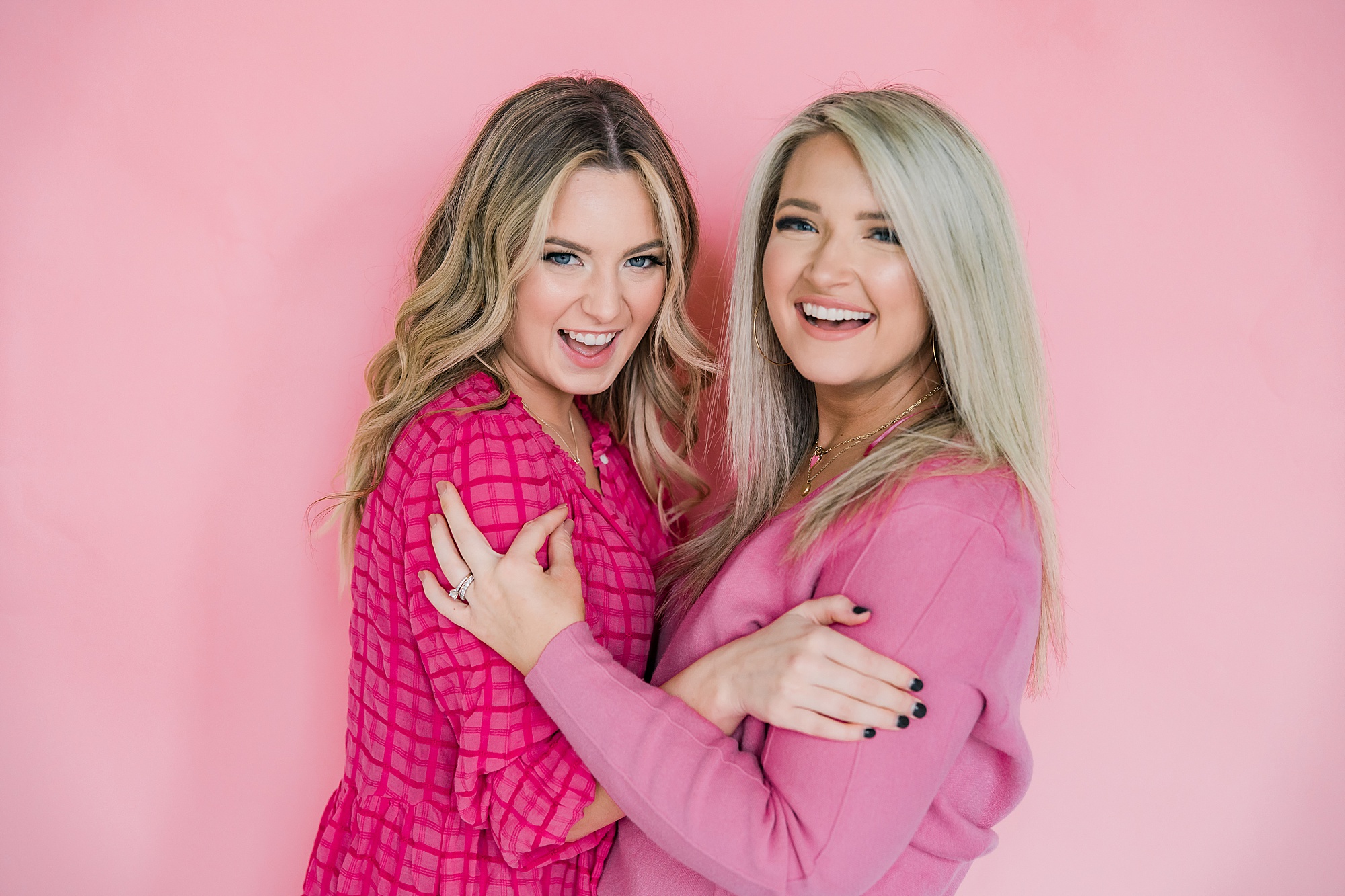 two women in pink sweaters hug against pink wall