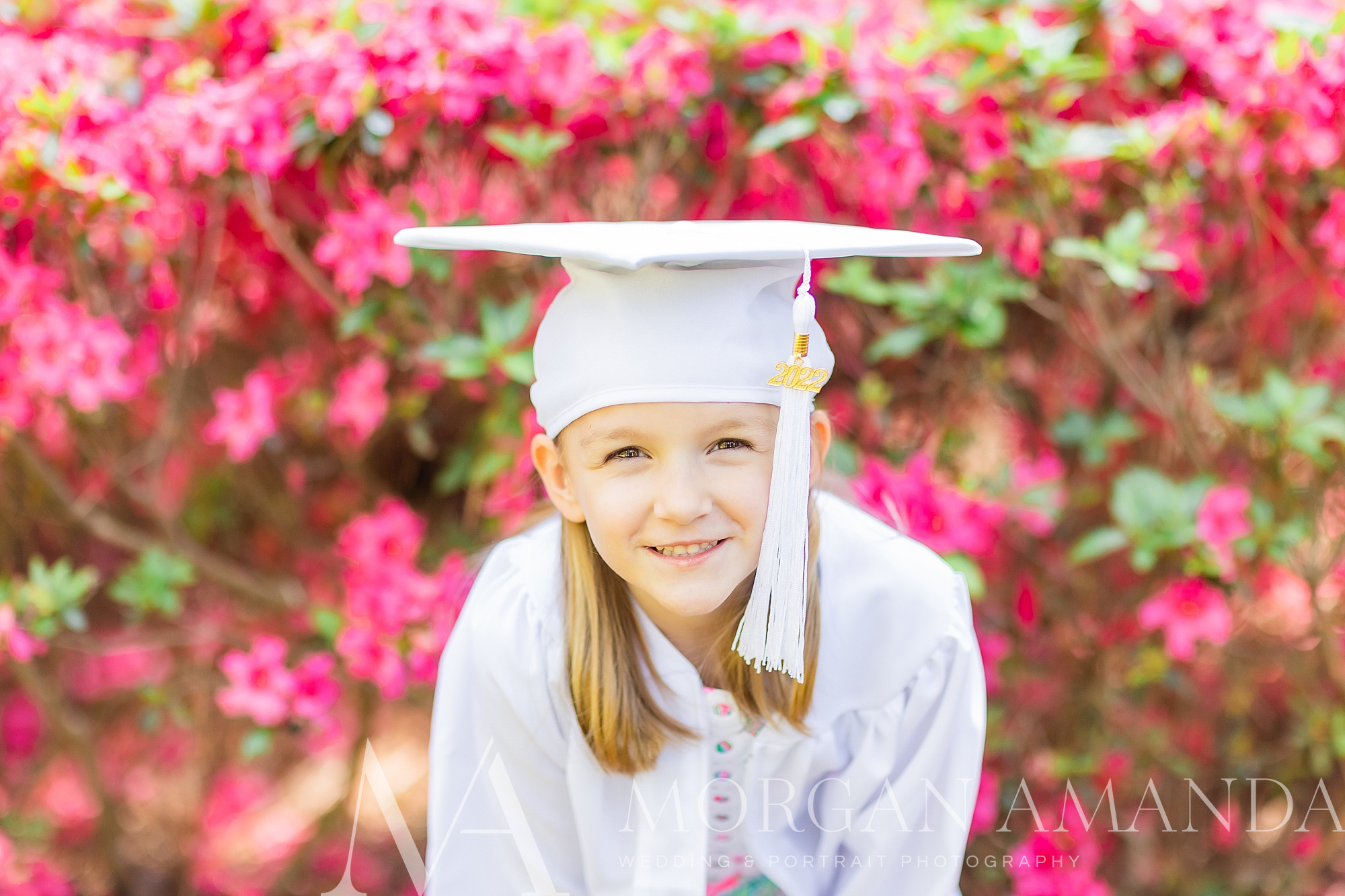 kindergartner poses by pink flowers in white cap and gown