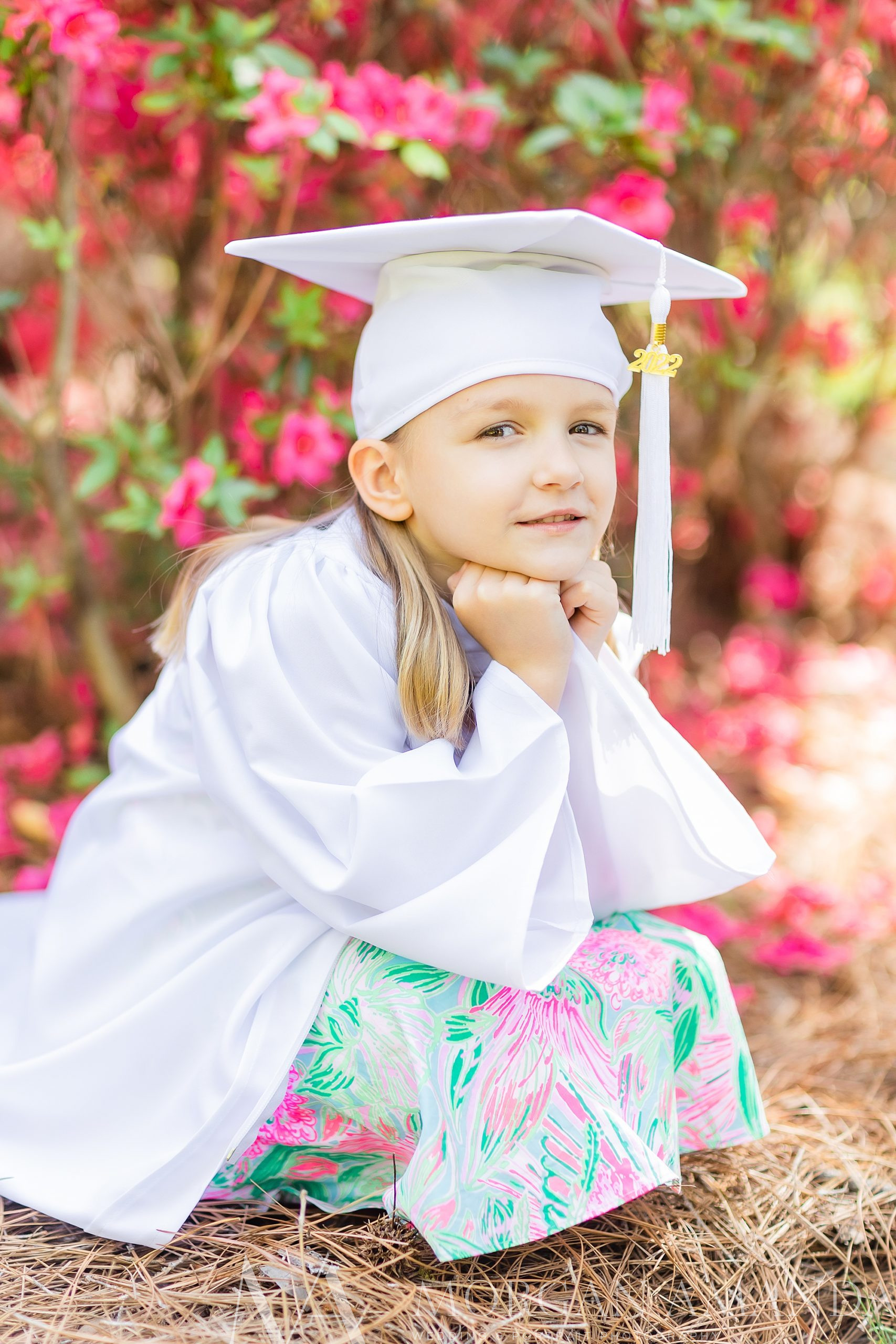 kindergartner sits with hands on knees in white cap and gown