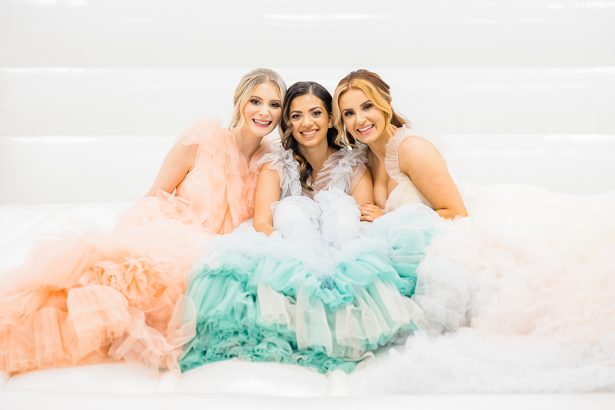 three women in pastel gowns sit on white bounce house