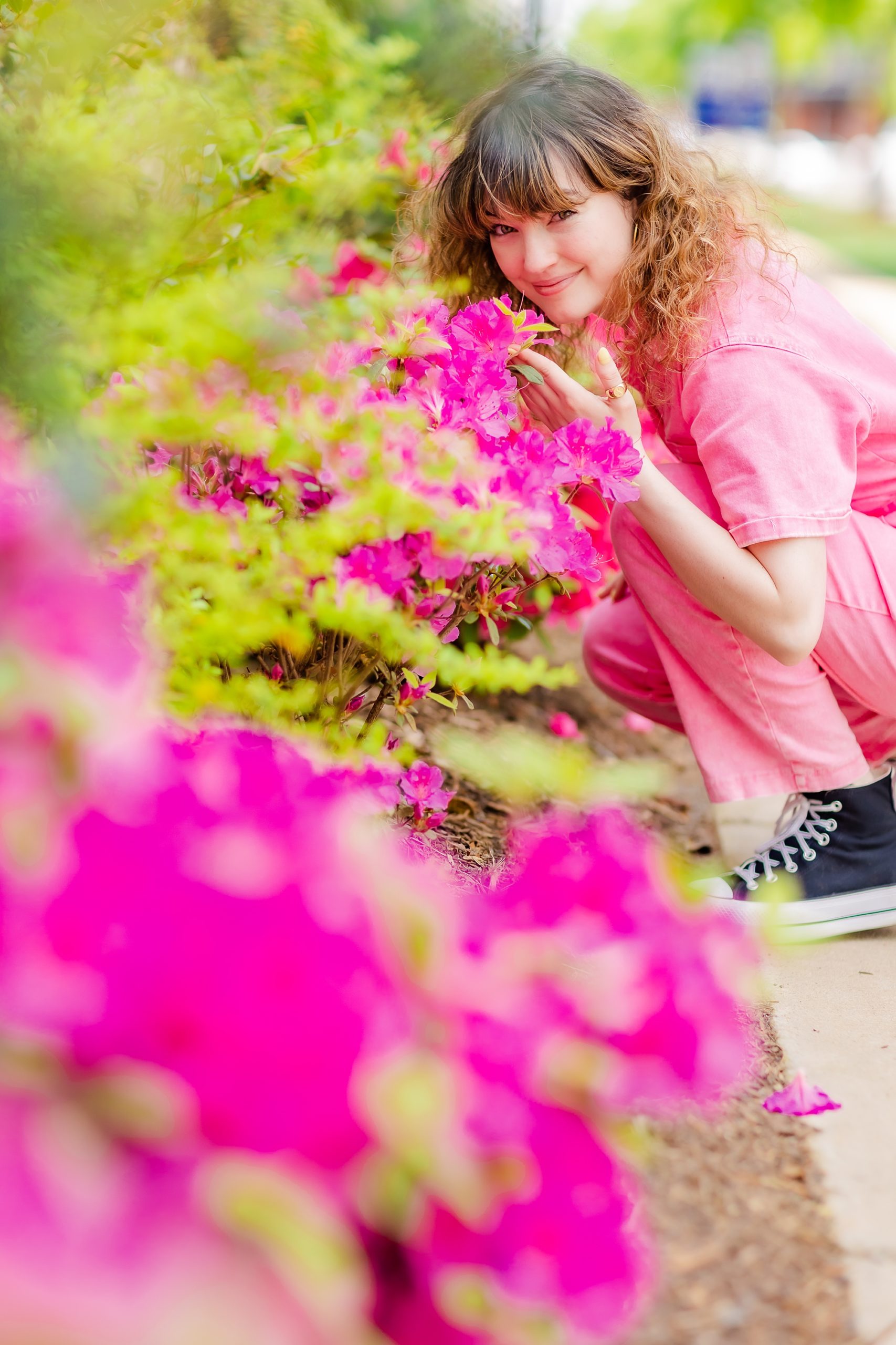 woman in pink jumpsuit leans to smell pink flowers 