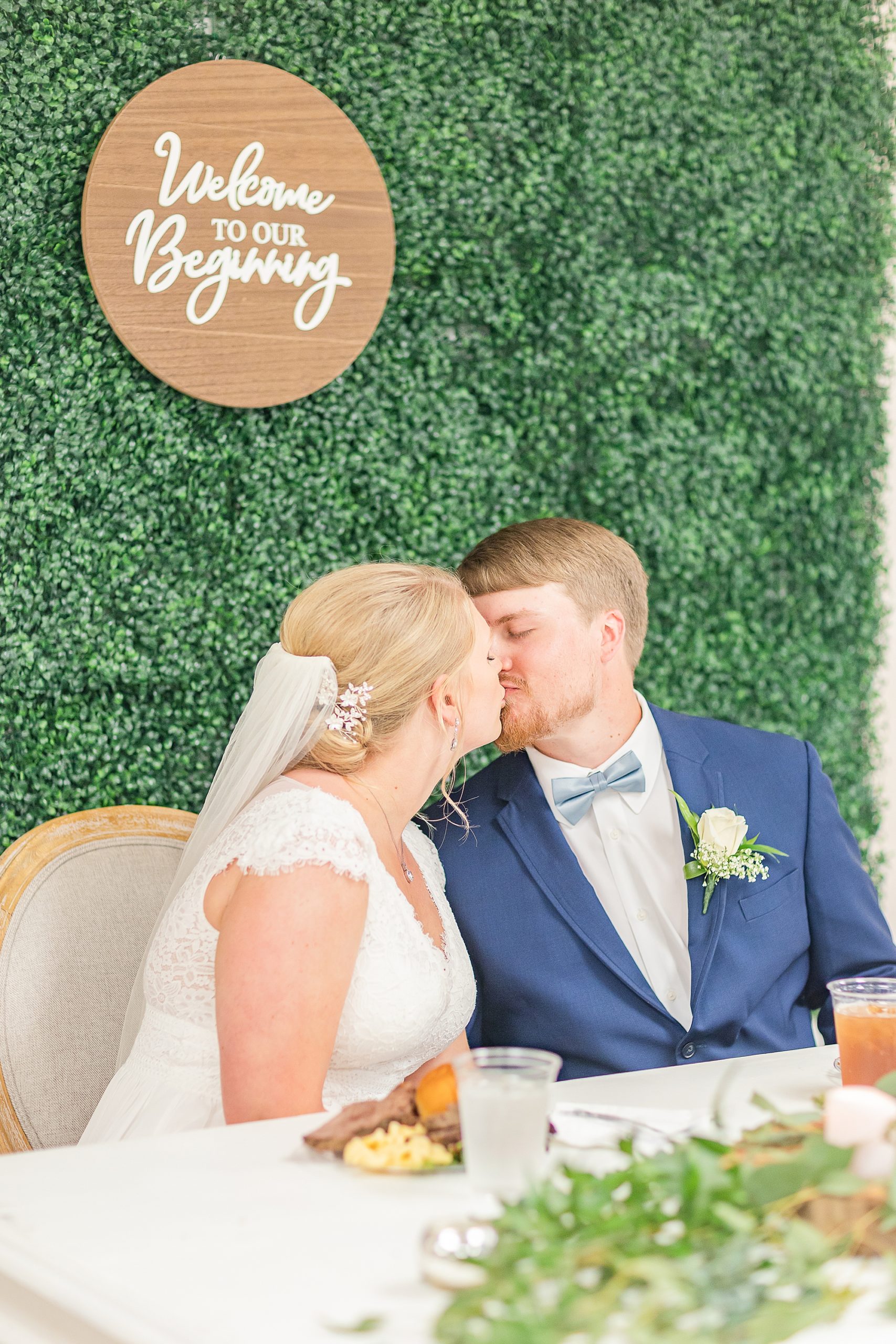 bride and groom kiss by wooden sign on greenery wall