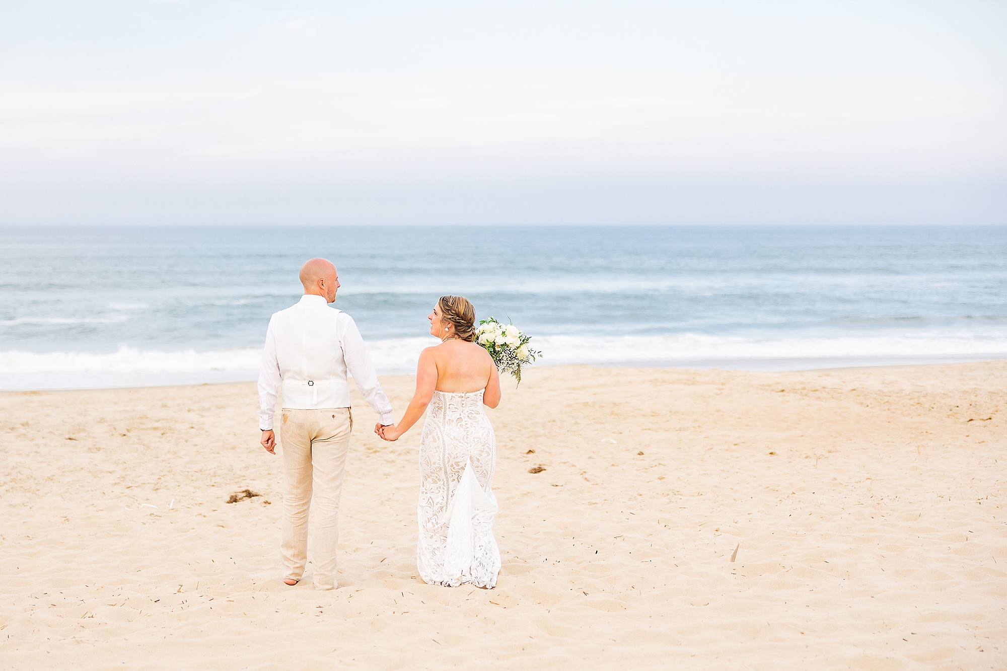 husband and wife hold hands walking across sand for OBX wedding photos