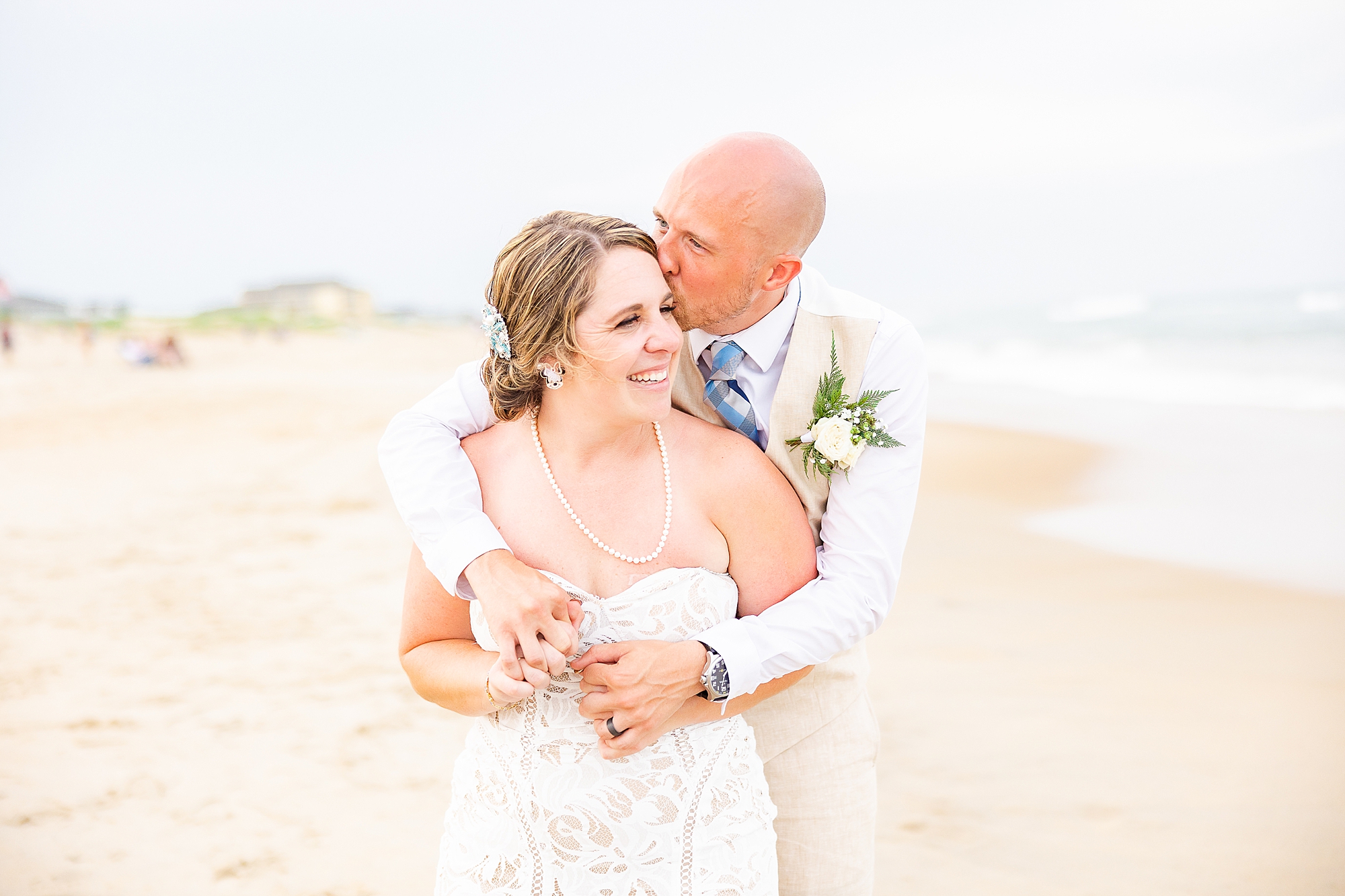 groom kisses bride's forehead from behind during wedding photos on beach