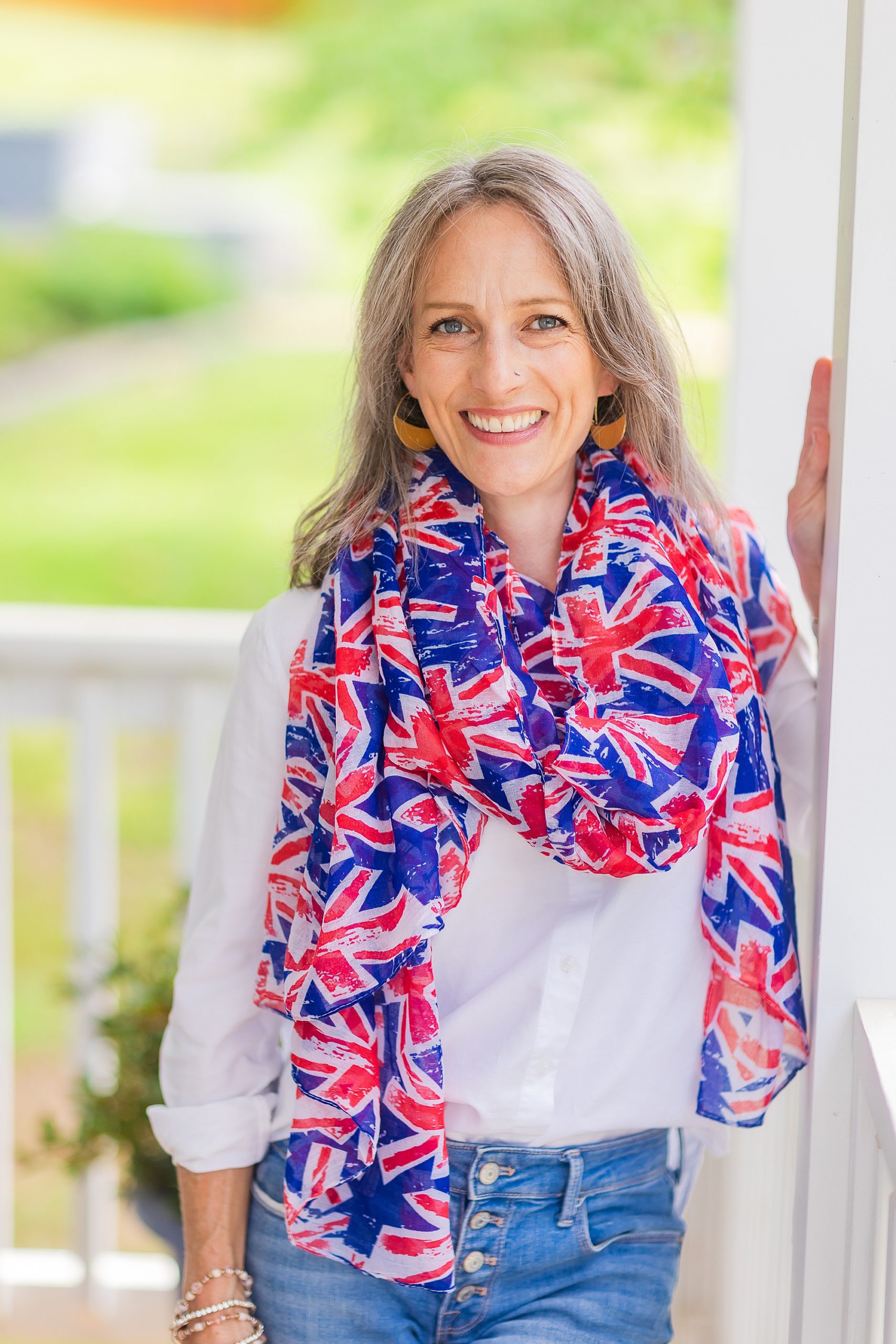 woman leans against railing of front porch in jeans and white shirt with UK flag scarf 
