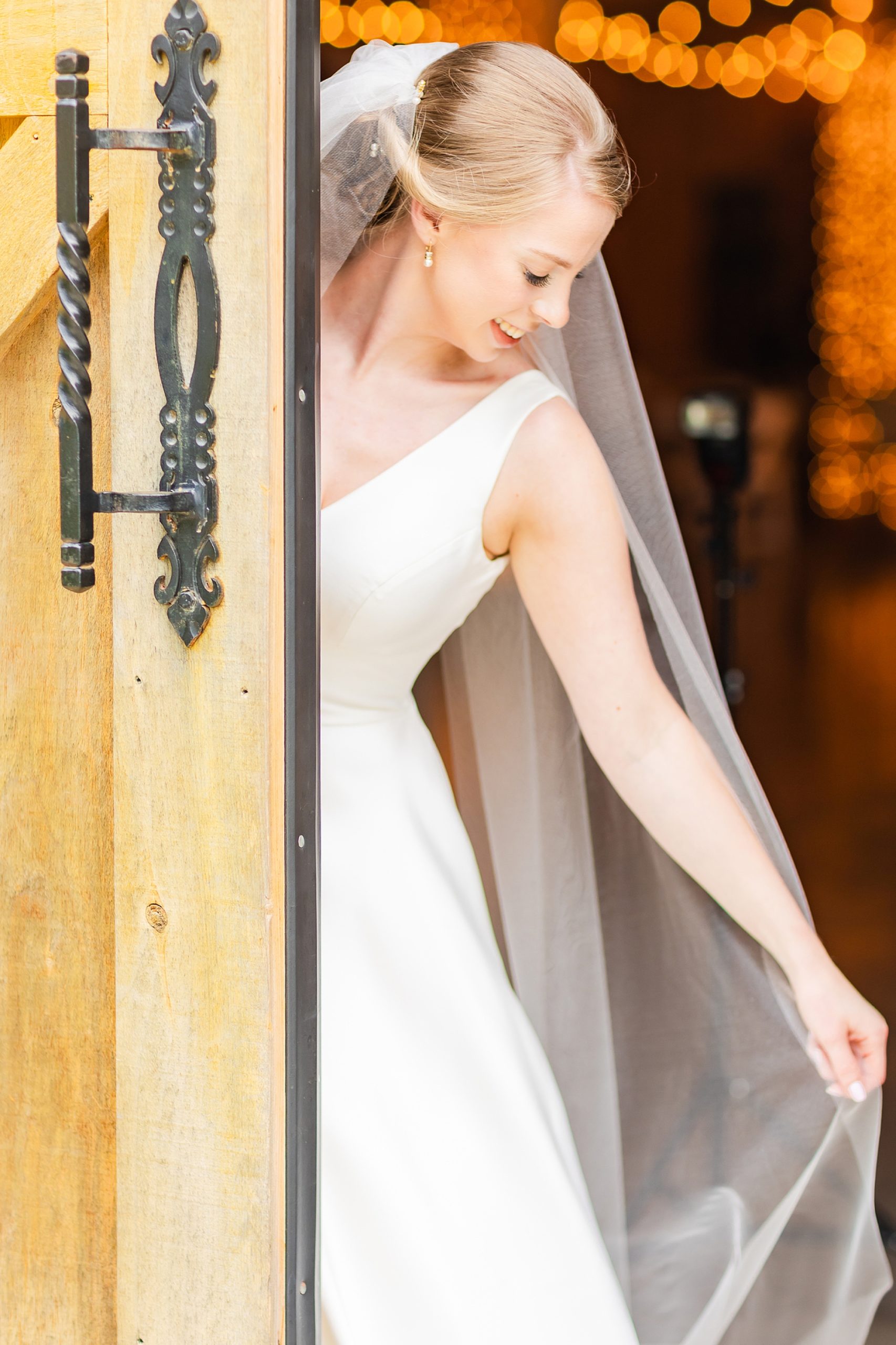 bride poses behind wooden door holding veil out beside her