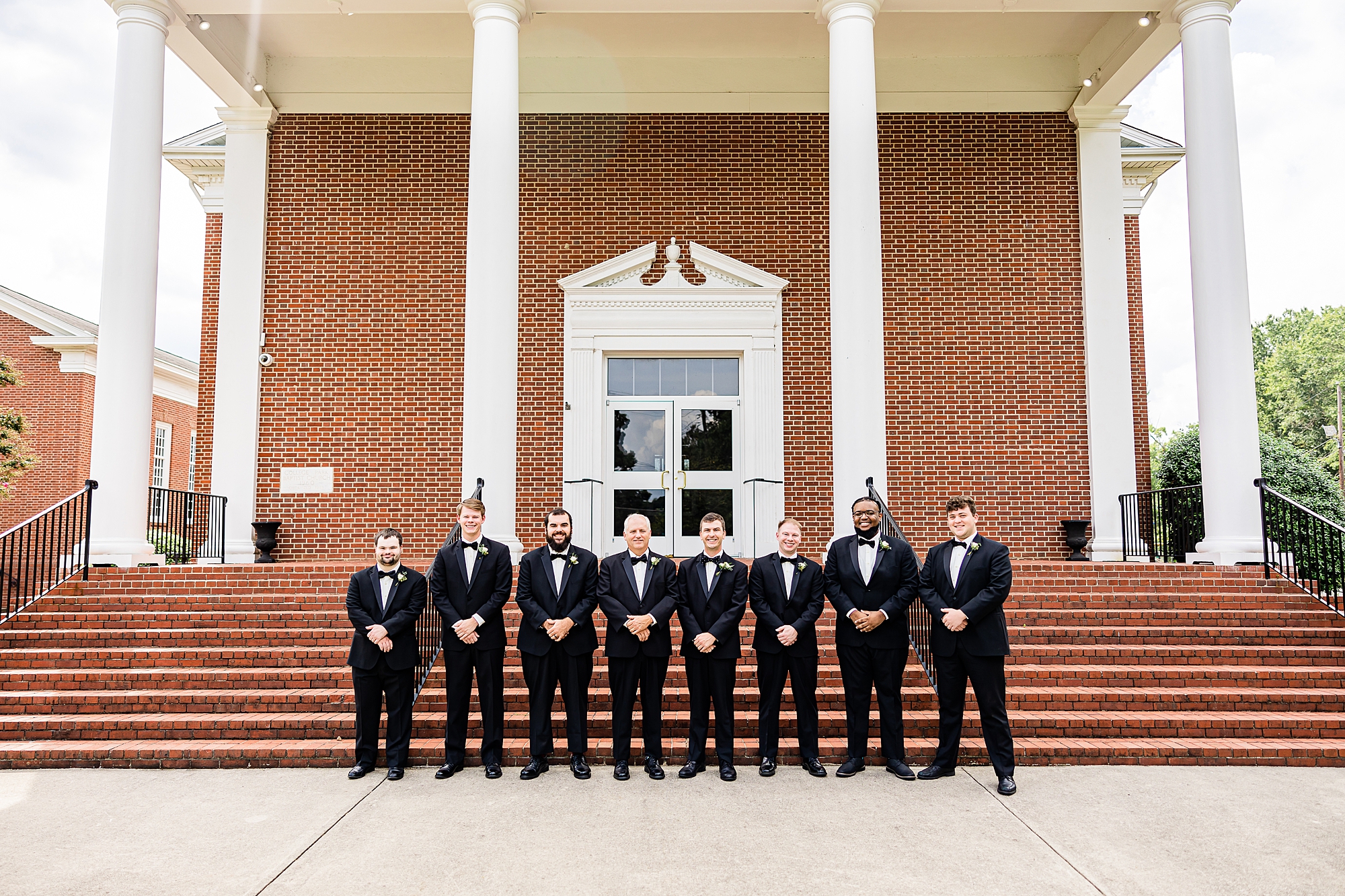groom and groomsmen stand outside College Park Baptist Church in black suits