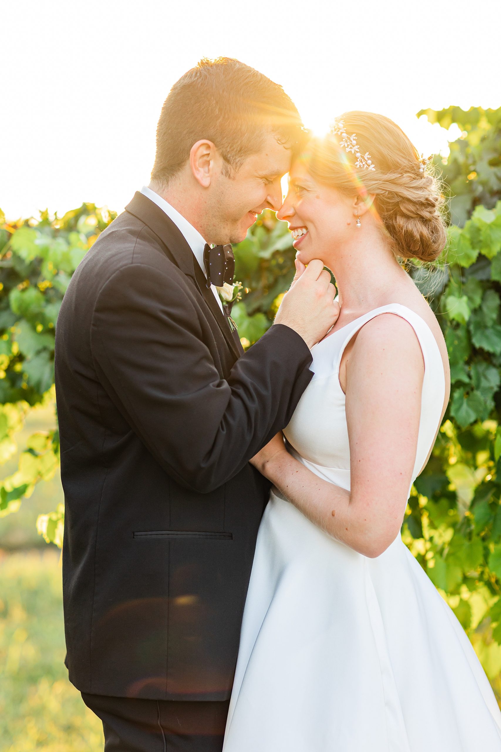groom holds bride's chin smiling at her between vines at Old Homeplace Vineyard