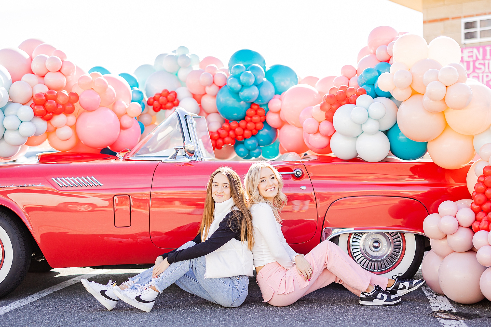 teen girls sit back to back by red car 