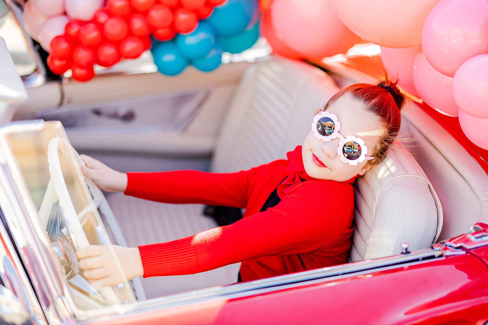 girl in read sweater pretends to drive car during Valentine’s Day photos